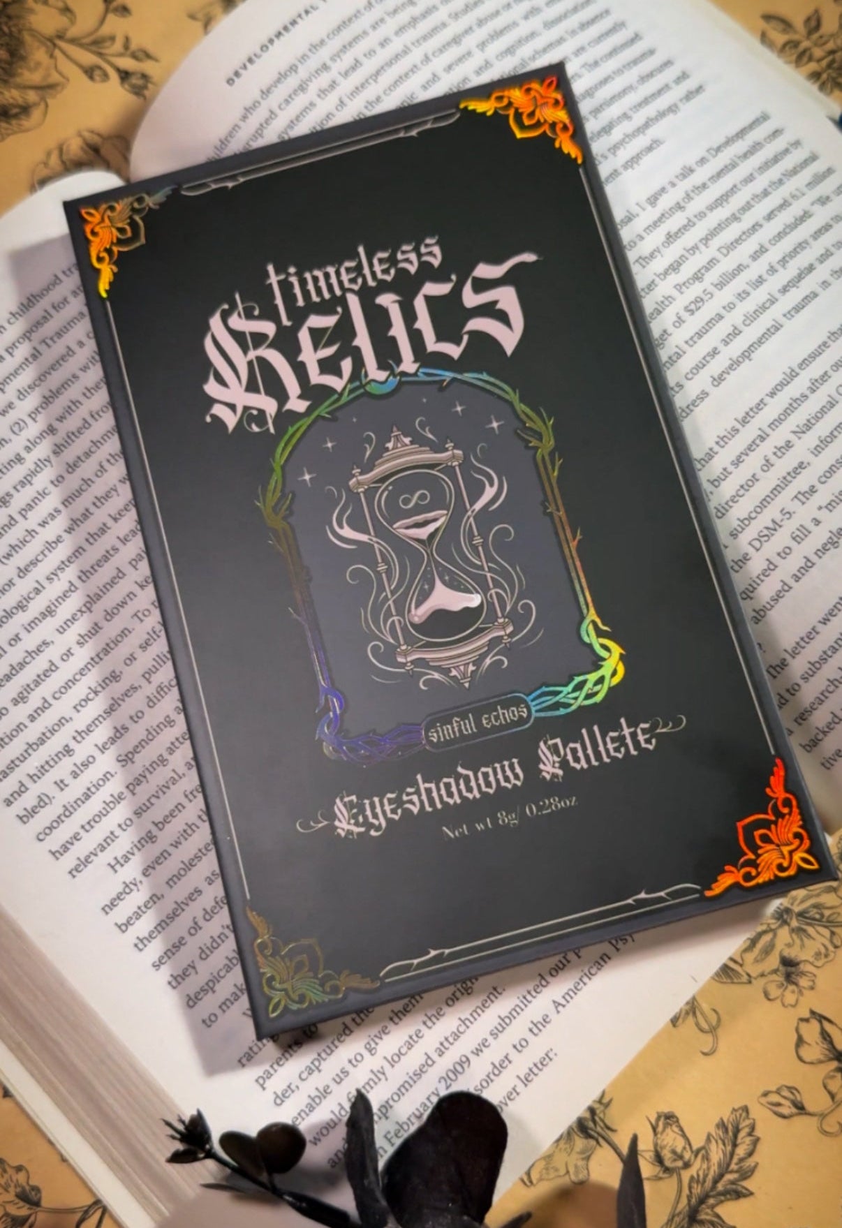 Timeless relics eyeshadow palette cover, with an hourgalss and gold holographic detailing, laying on top of an open book